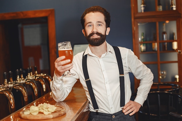 Man met snor en baard staat aan de bar en drinkt alcohol uit een glas.