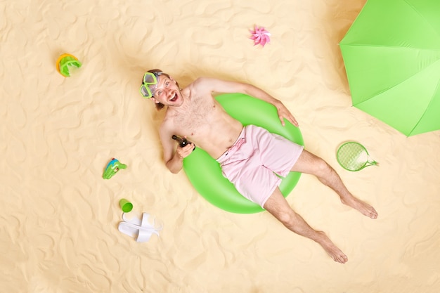 man poseert op groene zwemring met bier draagt korte broek snorkelmasker brengt vrije tijd door op het strand rust in de zomer kijkt vrolijk naar de camera