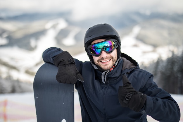 Man snowboarder staande op de top van de besneeuwde helling met snowboard, glimlachend naar de camera, duimen opdagen in winter skiresort.