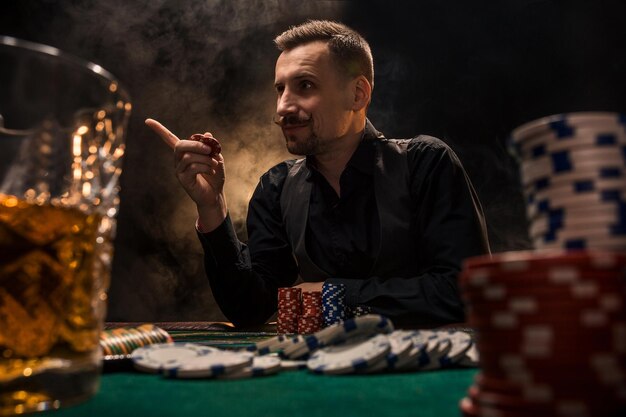 Man speelt poker met een sigaar en een whisky. Een man die alle fiches op tafel wint met dikke sigarettenrook. Het concept van de overwinning. Glas met whisky en stapel chips op de voorgrond