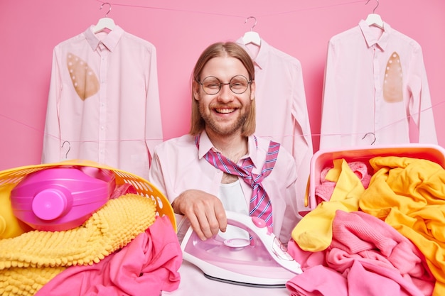 man strijkt kleren in waskamer staat vrolijk gebruikt elektris stroom ijzer draagt ronde bril shirt met stropdas om nek geïsoleerd op roze