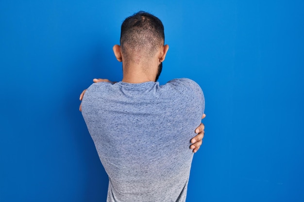 Man uit het midden-oosten met baard die over een blauwe achtergrond staat en zichzelf van achteren blij en positief omhelst. zelfliefde en zelfzorg