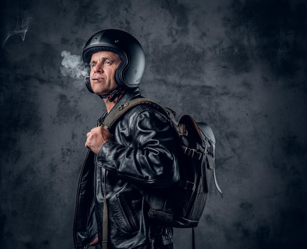 Man van middelbare leeftijd in motorhelm en leren jas met rugzak roken van een sigaret op grijze achtergrond.