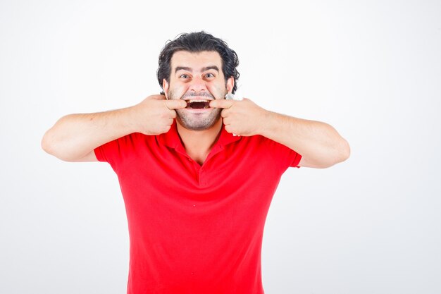 Man van middelbare leeftijd in rood t-shirt mond met handen opzij te trekken en grappig, vooraanzicht op zoek.