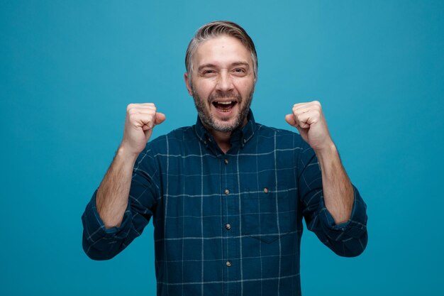 Man van middelbare leeftijd met grijs haar in donkere kleur shirt blij en opgewonden gebalde vuisten verheugd over zijn succes staande over blauwe achtergrond