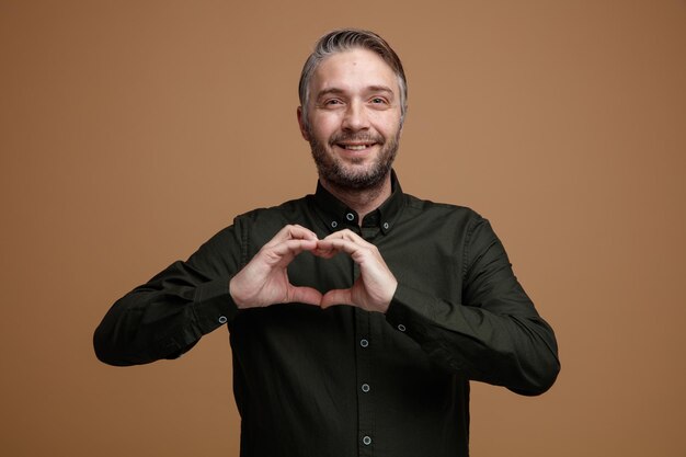 Gratis foto man van middelbare leeftijd met grijs haar in donkere kleur shirt kijken camera blij en positief hart gebaar maken met handen over zijn borst glimlachend staande over bruine achtergrond