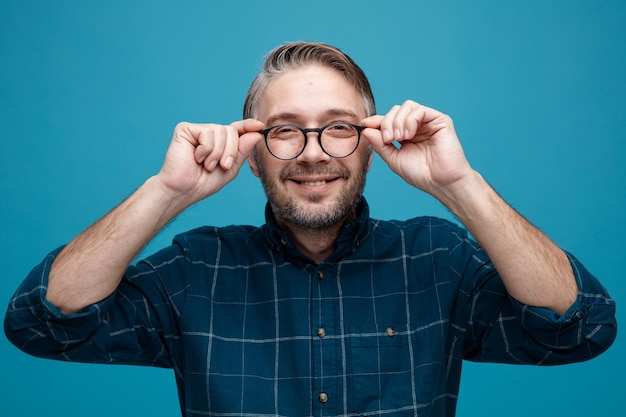 Man van middelbare leeftijd met grijs haar in donkere kleur shirt met bril kijkend naar camera glimlachend vrolijk gelukkig en positief staande over blauwe achtergrond