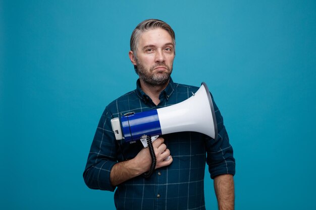 Man van middelbare leeftijd met grijs haar in donkere kleur shirt met megafoon kijkend naar camera met sceptische glimlach op gezicht staande over blauwe achtergrond