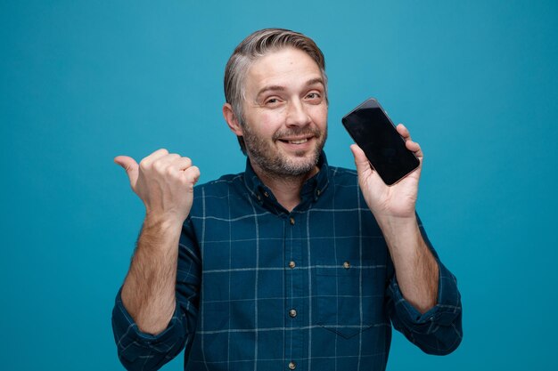 Man van middelbare leeftijd met grijs haar in donkere kleur shirt met smartphone kijken camera glimlachend wijzend met duim naar de zijkant staande over blauwe achtergrond