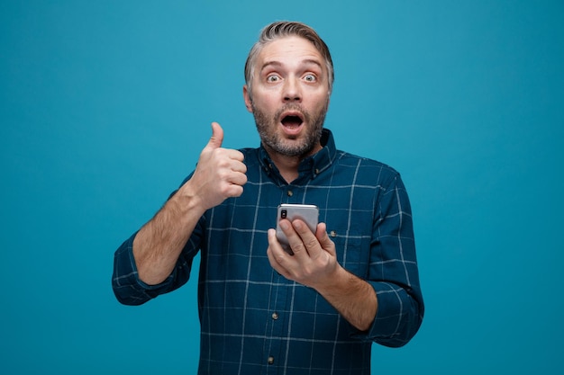 Man van middelbare leeftijd met grijs haar in donkere kleur shirt met smartphone met duim omhoog kijkend naar camera verbaasd en verrast staande over blauwe achtergrond