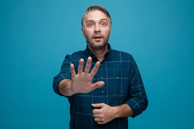 Man van middelbare leeftijd met grijs haar in een donker gekleurd hemd dat naar de camera kijkt, bezorgd een stopgebaar maakt met de hand die over een blauwe achtergrond staat