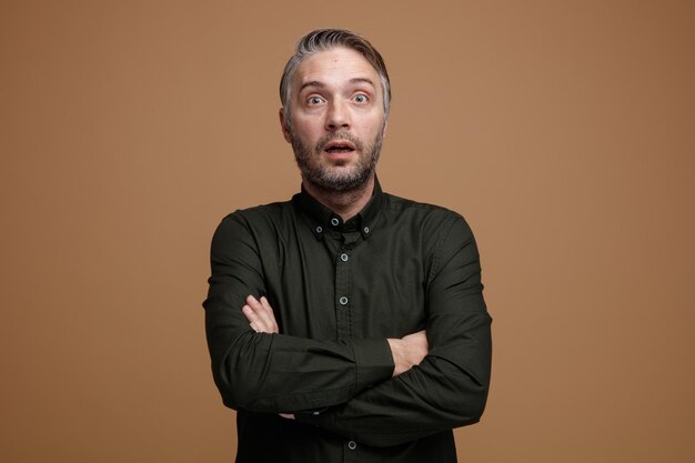 Man van middelbare leeftijd met grijs haar in een donker gekleurd hemd dat naar de camera kijkt verbaasd en verrast terwijl hij zijn handen op zijn borst kruist en over een bruine achtergrond staat