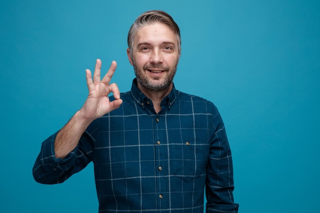 Gratis foto man van middelbare leeftijd met grijs haar in een donker gekleurd hemd die naar de camera kijkt, blij en positief glimlacht en een ok teken toont dat over een blauwe achtergrond staat