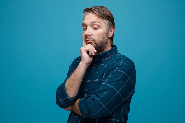 Man van middelbare leeftijd met grijs haar in een donker gekleurd hemd die opzij kijkt en denkt terwijl hij zijn wang aanraakt die over een blauwe achtergrond staat