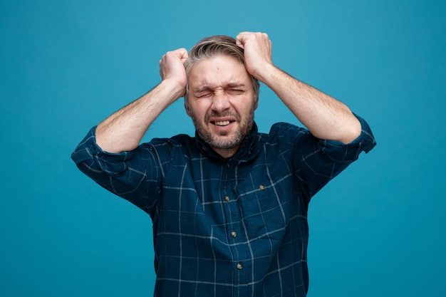 Gratis foto man van middelbare leeftijd met grijs haar in een donker gekleurd hemd, gek gek die zijn haar gestrest en nerveus over een blauwe achtergrond trekt