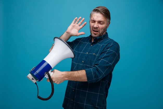 Man van middelbare leeftijd met grijs haar in een donker gekleurd hemd met een megafoon die ernaar kijkt omdat hij ontevreden over een blauwe achtergrond staat