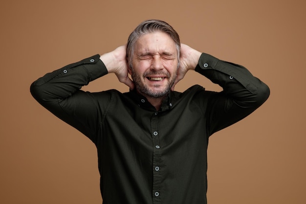 Gratis foto man van middelbare leeftijd met grijs haar in een donkere kleur shirt blij en opgewonden hand in hand achter zijn hoofd staande over bruine achtergrond