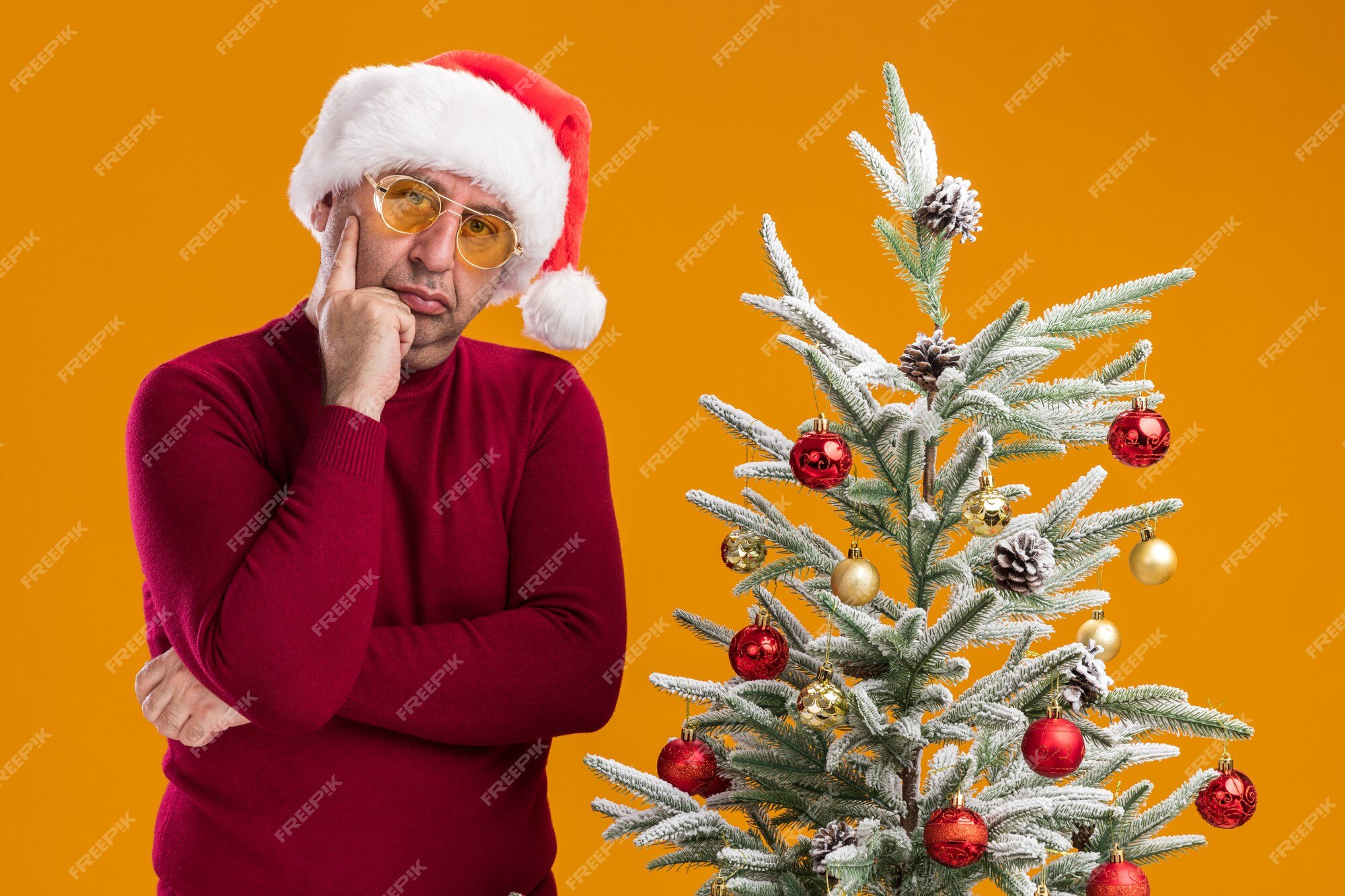 Man van middelbare leeftijd met kerst-kerstmuts in donkerrode en gele bril camera kijken verbaasd staande naast een kerstboom over oranje achtergrond | Gratis Foto