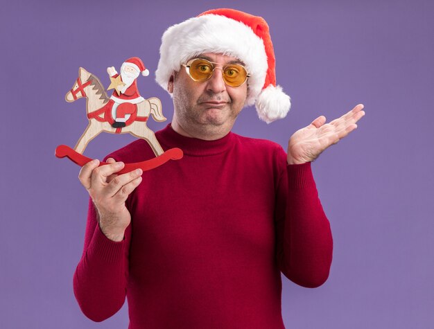 Man van middelbare leeftijd met kerst kerstmuts in gele bril met kerst speelgoed kijken camera verward en ontevreden staande over paarse achtergrond