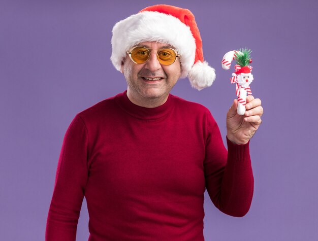 Man van middelbare leeftijd met kerst kerstmuts in gele glazen met kerst speelgoed kijken camera met glimlach op gezicht staande over paarse achtergrond