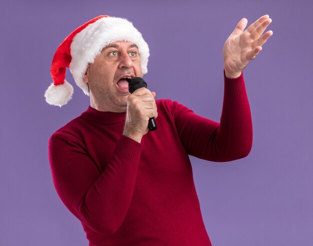 Man van middelbare leeftijd met kerst kerstmuts met microfoon zingen blij en opgewonden staande over paarse achtergrond