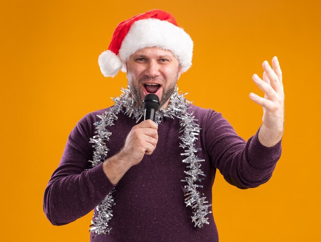 Man van middelbare leeftijd met kerstmuts en klatergoud guirlande rond de nek met microfoon kijken camera houden hand in lucht zingen geïsoleerd op oranje achtergrond