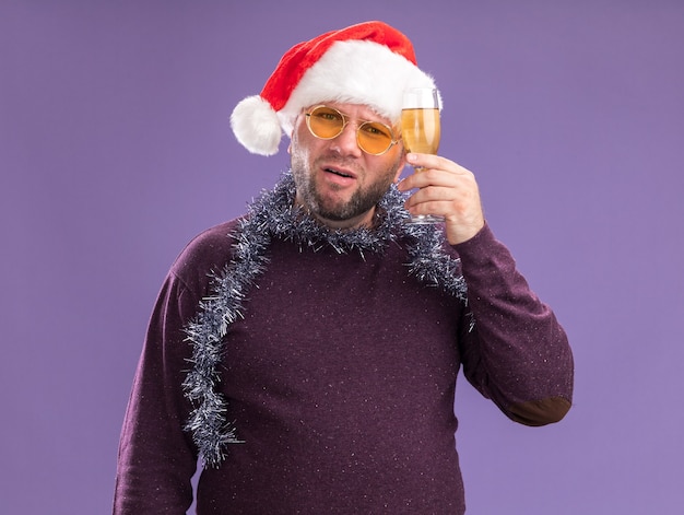 Man van middelbare leeftijd met kerstmuts en klatergoud slinger rond de nek met een glas champagne geïsoleerd op paarse muur