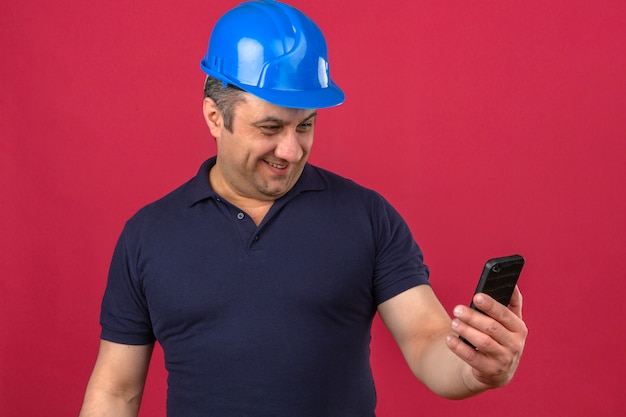 Man van middelbare leeftijd met poloshirt en veiligheidshelm kijken naar het scherm van de mobiele telefoon met een grote glimlach op het gezicht over geïsoleerde roze muur