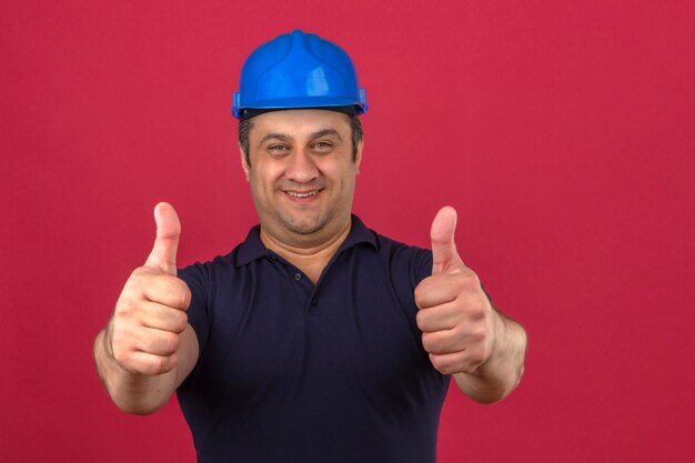 Man van middelbare leeftijd met poloshirt en veiligheidshelm met een glimlach op het gezicht en duimen opdagen over geïsoleerde roze muur