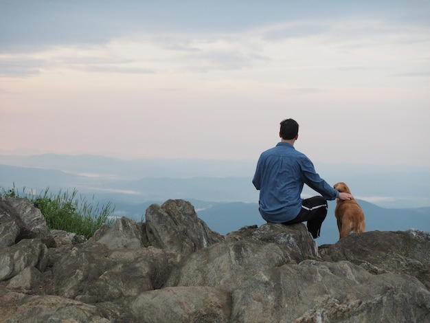 Man zittend op de rots en een hond aaien, omringd door bergen onder een bewolkte hemel