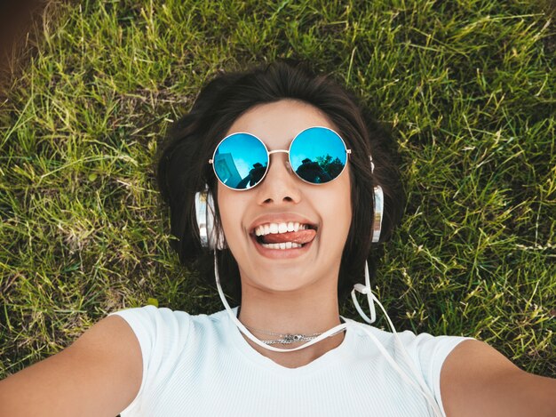 Manierportret van jonge modieuze hipstervrouw die op het gras in het park liggen. Het meisje draagt trendy uitrusting Glimlachend model makend selfie. Vrouw luisteren naar muziek via een koptelefoon