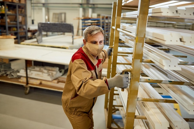 Mannelijke arbeider met gezichtsmasker bewegend rek met verwerkt hout bij timmerwerkplaats