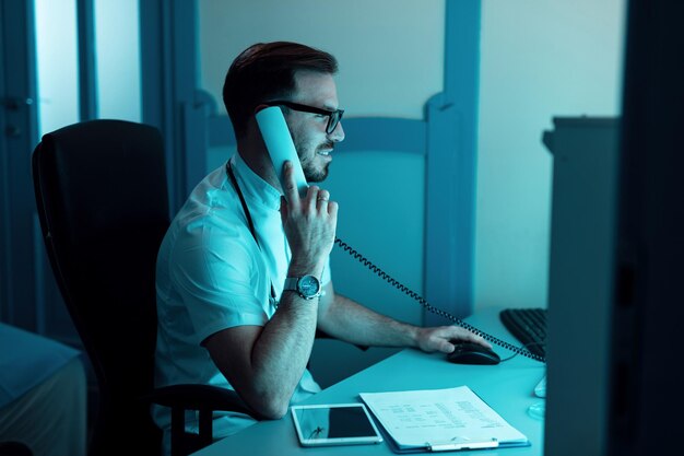 Mannelijke arts die aan een computer werkt en via de telefoon in het ziekenhuis communiceert