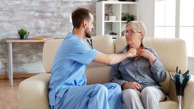 Mannelijke arts die zijn stethoscoop opzet en luistert naar de hartslag van de oude vrouw in het verpleeghuis