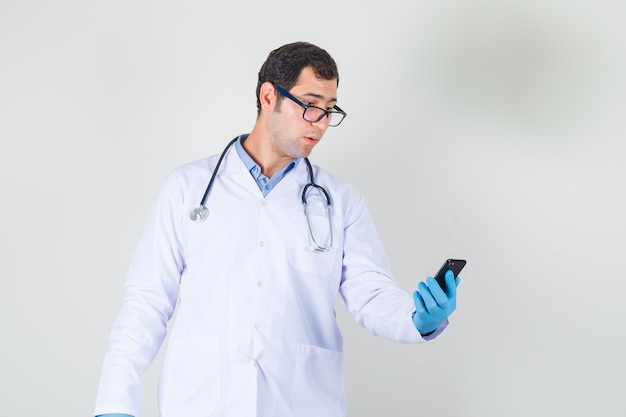 Mannelijke arts houden en kijken naar smartphone in witte jas, handschoenen, glazen vooraanzicht.
