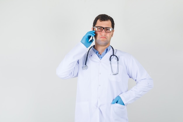 Mannelijke arts in witte jas, handschoenen, bril praten over de telefoon met hand in de zak en ontevreden op zoek