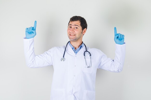Mannelijke arts in witte jas, handschoenen vingers omhoog en op zoek vrolijk