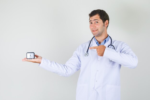 Mannelijke arts in witte jas wijzende vinger op klok en op zoek vrolijk