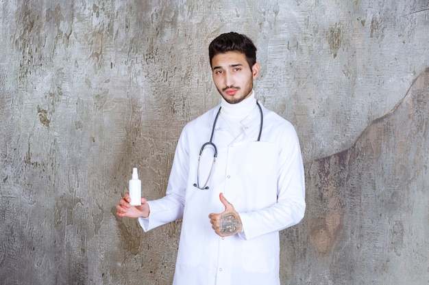 Mannelijke arts met een stethoscoop die een witte fles van het handdesinfecterend middel houdt en positief handteken toont.