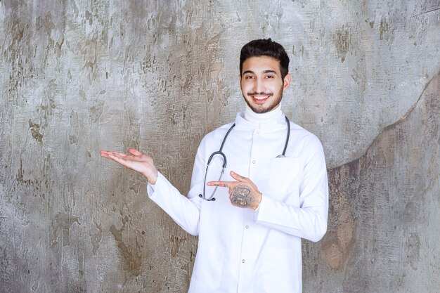 Mannelijke arts met een stethoscoop die zich op betonnen muur bevindt en iets aan de linkerkant laat zien.