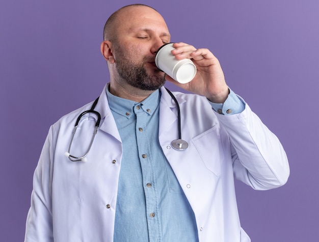 Mannelijke arts van middelbare leeftijd met een medisch gewaad en een stethoscoop die koffie drinkt uit een plastic koffiekopje met gesloten ogen geïsoleerd op een paarse muur