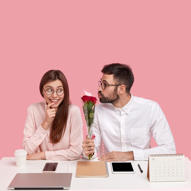 Mannelijke baas werd verliefd op jonge mooie collega, geeft prachtige rode rozen, vouwt de lippen voor het maken van een kus, gelukkige dame ontvangt compliment en bloemen, zit op het bureaublad in kantoor tegen roze muur