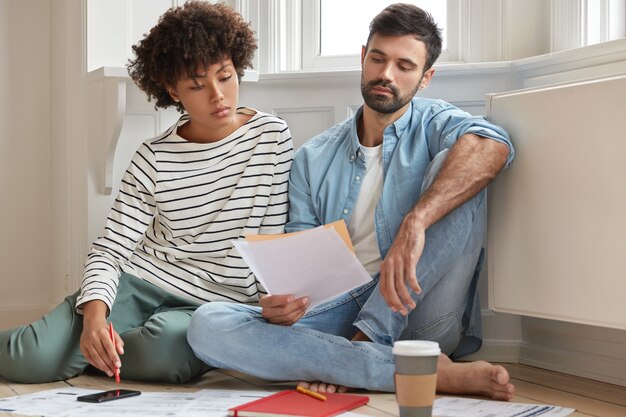 Mannelijke bebaarde accountant en zijn secretaresse werken samen in een modern appartement, poseren op een houten vloer en bespreken financieel verslag, hebben een serieuze blik in kranten, studeren analyses, voelen zich thuis op hun gemak