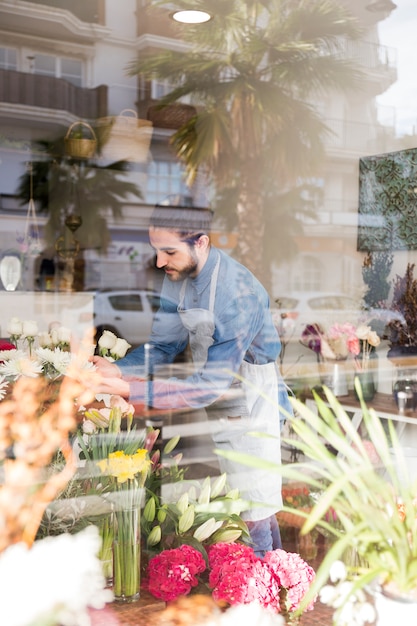 Gratis foto mannelijke bloemist die de bloemen schikt die door het glas in de bloemwinkel worden gezien