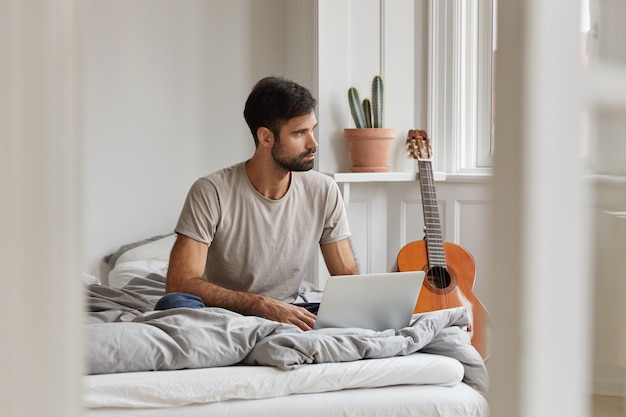 Mannelijke blogger heeft een doordachte uitdrukking, geniet van vrije tijd met laptop, zit op een comfortabel bed