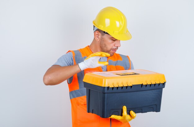 Mannelijke bouwer die plastic gereedschapskist in uniform, helm, handschoenen, vooraanzicht houdt.