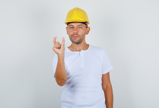 Mannelijke bouwer doet klein formaat bord met vingers in wit t-shirt, veiligheidshelm vooraanzicht.