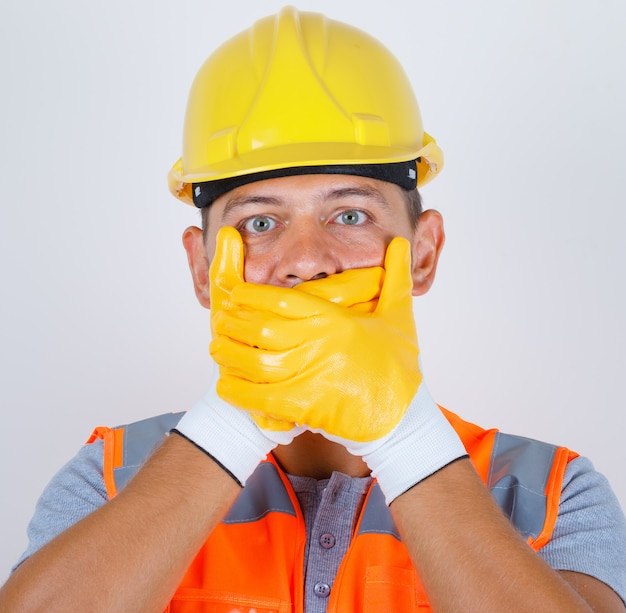 Gratis foto mannelijke bouwer in uniform, helm, handschoenen die mond bedekken met handen voor fout en op zoek geschokt, vooraanzicht.