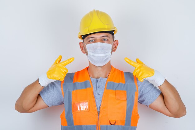 Mannelijke bouwer wijst zijn medisch masker in uniform, helm, handschoenen, vooraanzicht