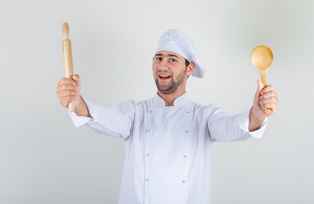 Mannelijke chef-kok die houten lepel en deegroller in wit uniform houdt en vrolijk kijkt